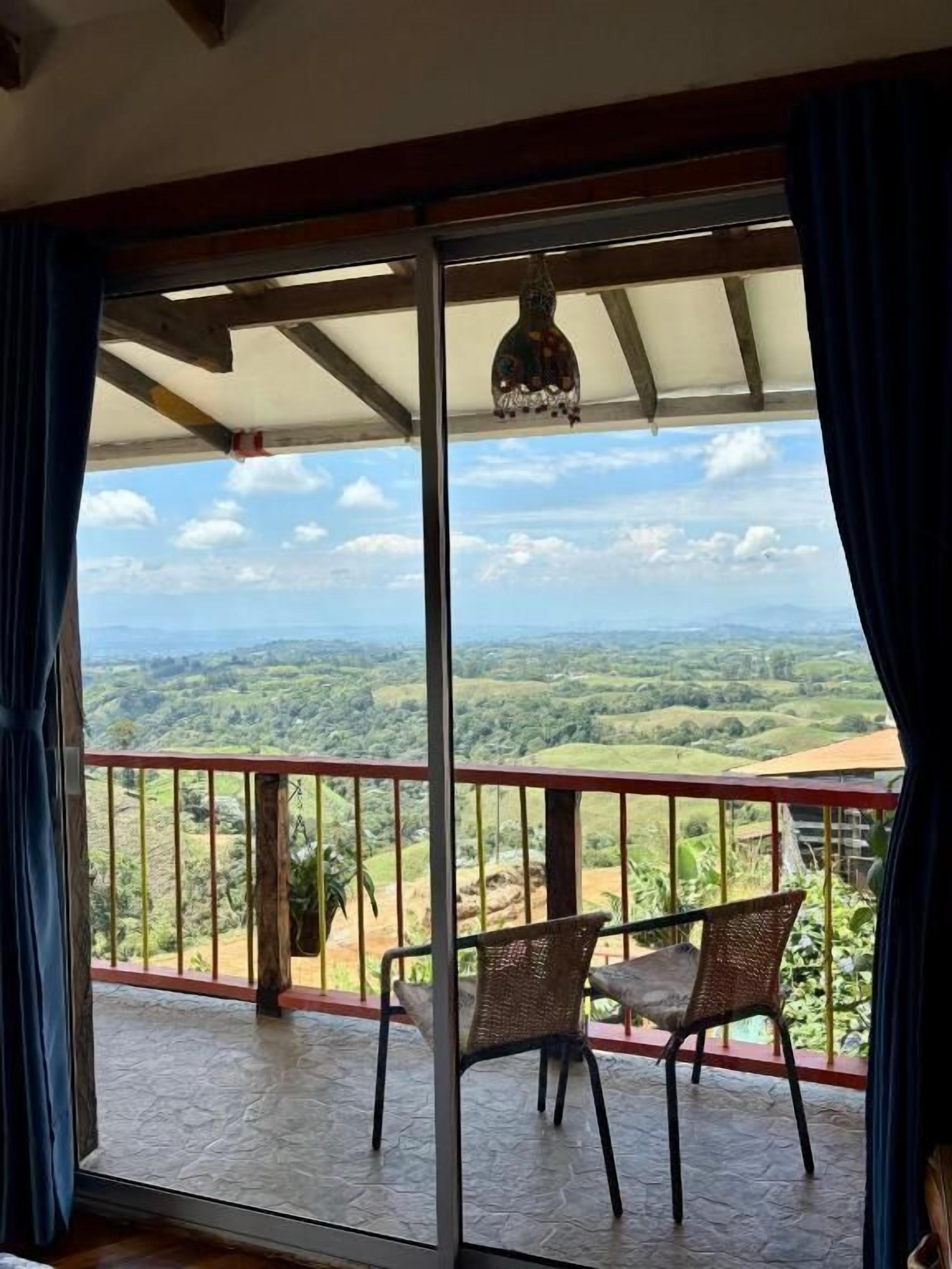 Un balcón con vistas a un paisaje verde, un adorno colgante y dos sillas de mimbre.