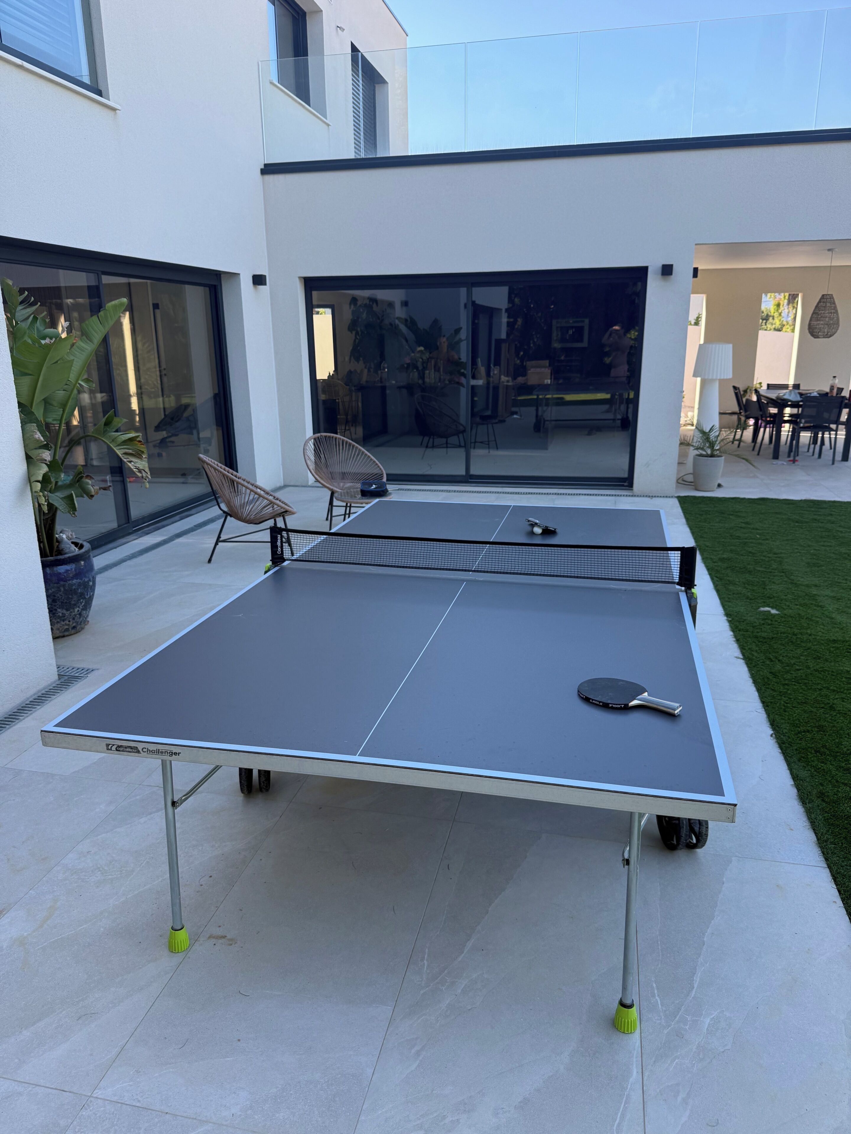 An outdoor ping pong table with a paddle, chairs, and a potted plant.