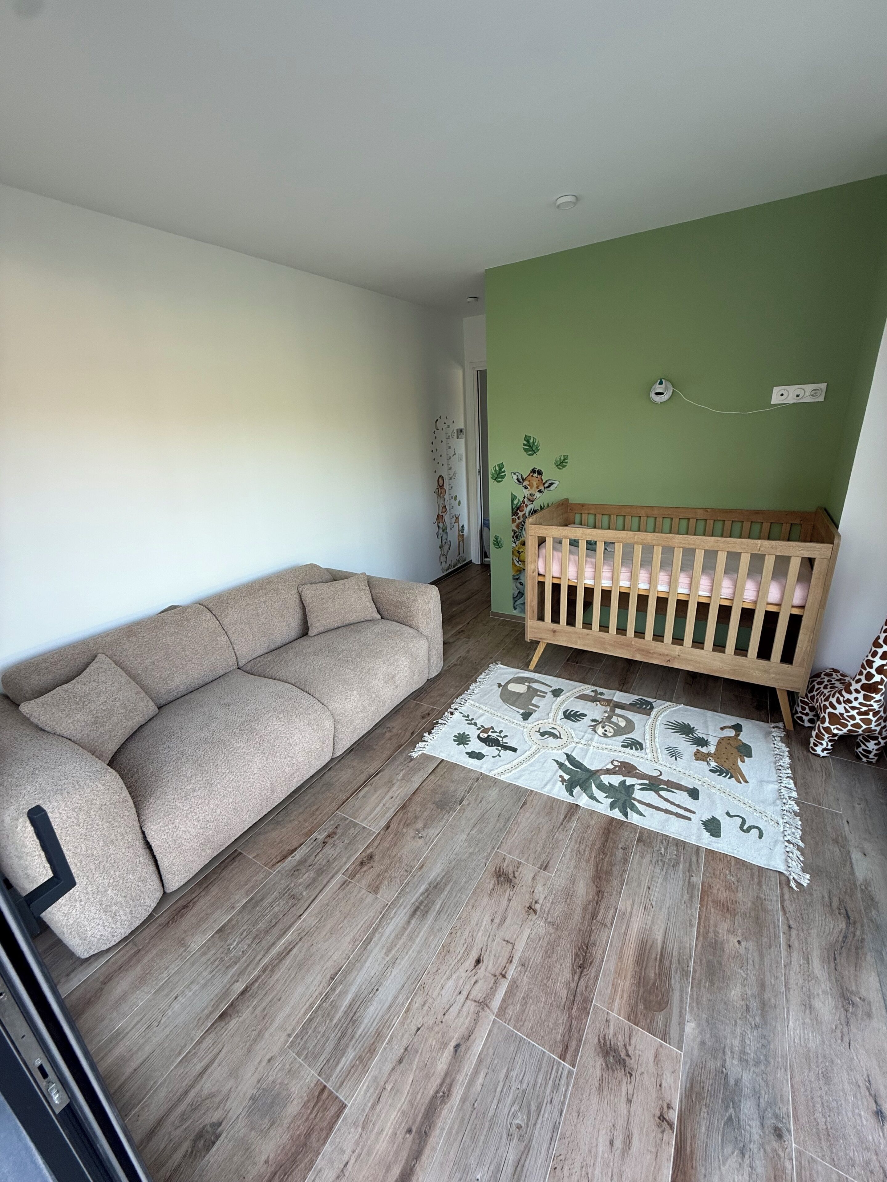 Een kamer met een beige bank, een houten kinderbedje, een kamerkleed met dessin en een groene muur met girafsticker.