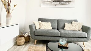 Ein graues Sofa mit weißen Kissen, ein Glastisch, ein gewebter Korb und eine Vase mit Trockenpflanzen.