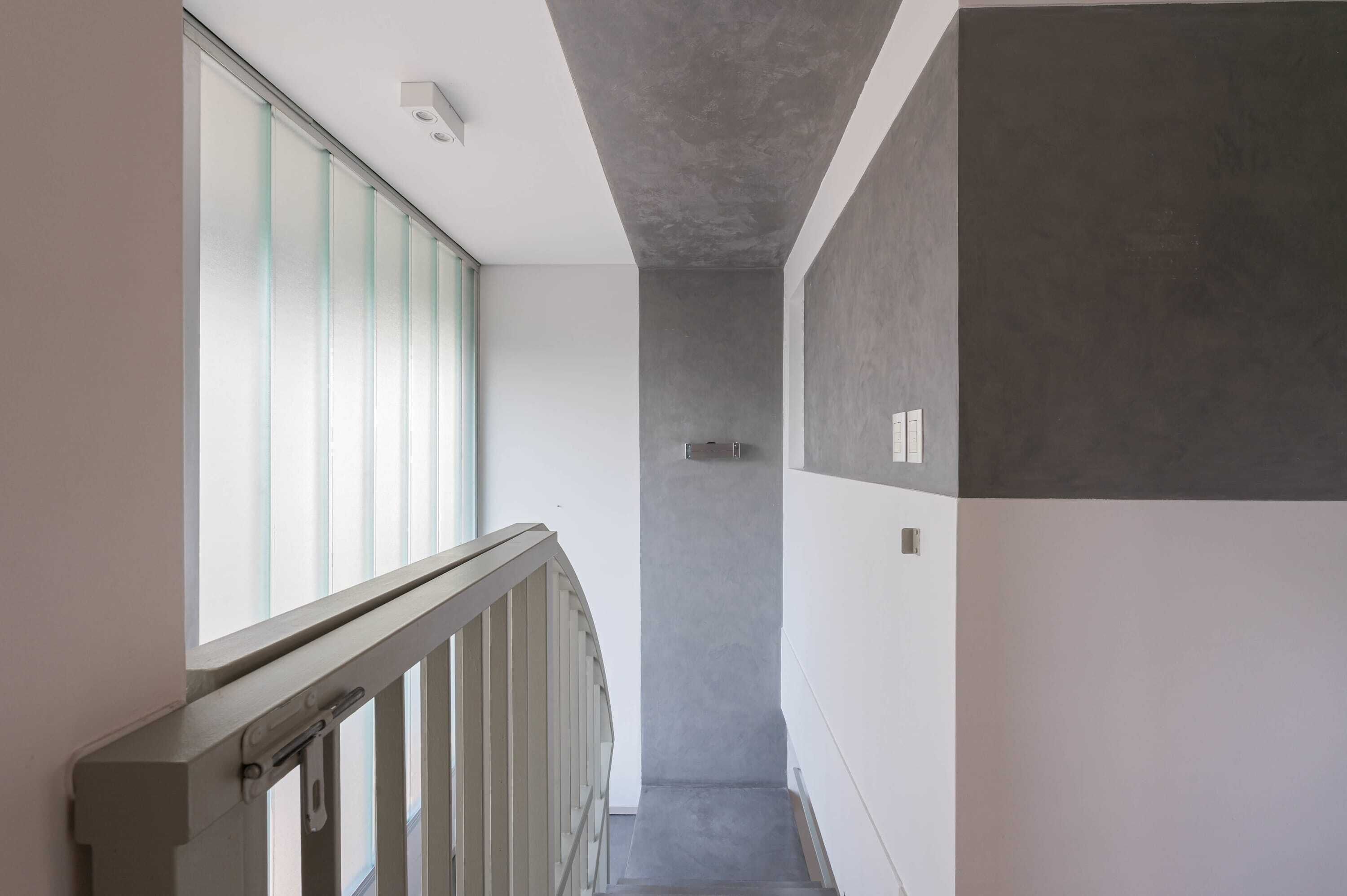 Un couloir étroit avec un escalier, une fenêtre avec des stores et un mur gris.