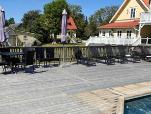 A wooden deck with lounge chairs and umbrellas, a pool, and a building with a red roof in the background. - 4 Star Holiday Home in Gotlands Tofta (Gotlands Tofta)