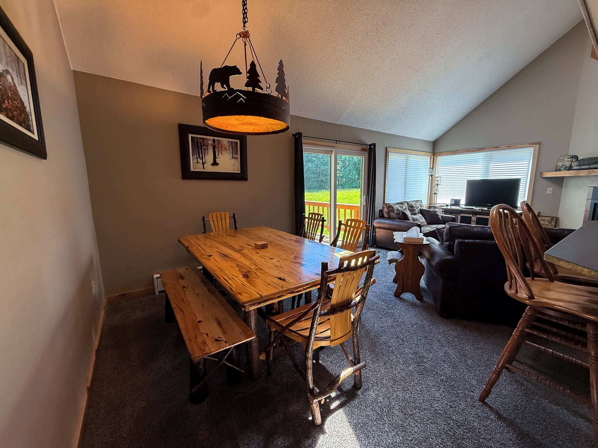 A dining area with a wooden table and chairs, a hanging light fixture, and a view of the outdoors through a sliding glass door.