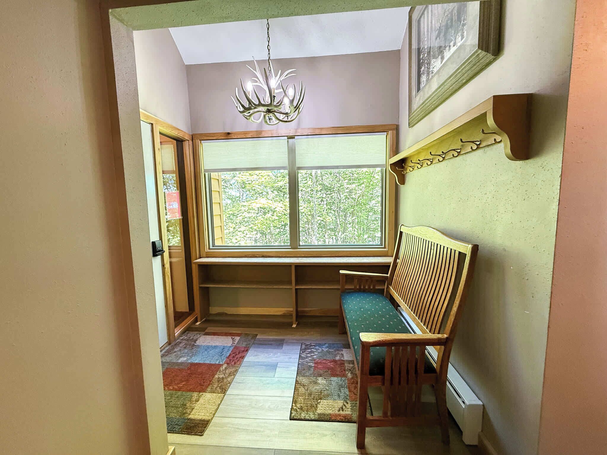 A corner room with a bench, a window with blinds, a chandelier, and a patterned rug.