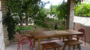 An outdoor patio with a wooden table and chairs, surrounded by greenery and a stone wall. - Apartments Snjezana Carev - Studio Apartment With Balcony (Tisno)