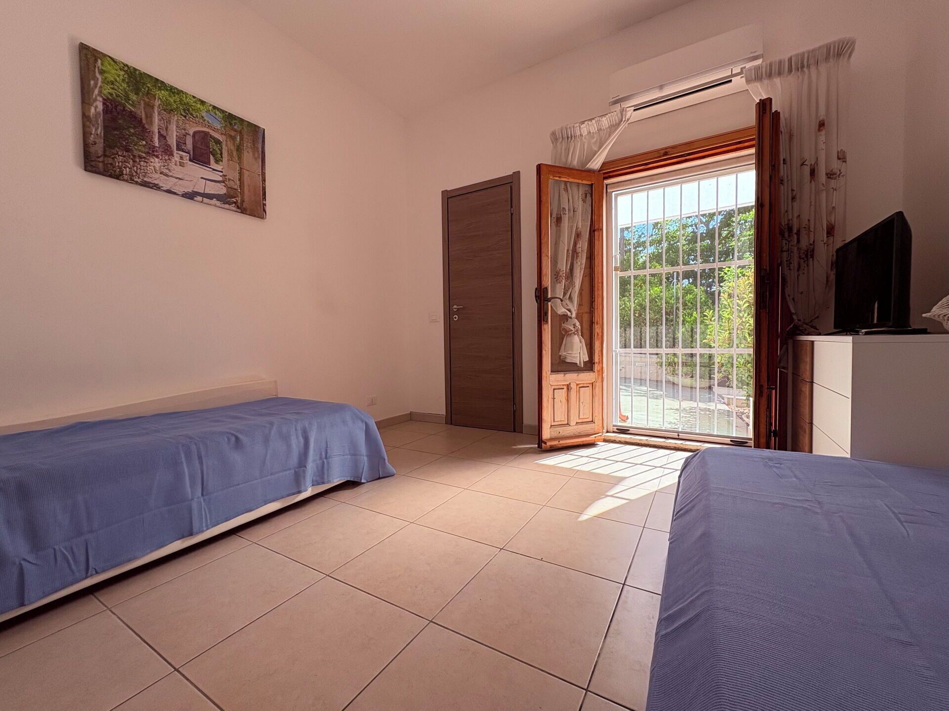 Ein Zimmer mit zwei Betten, einem Gemälde, einem Fernseher und einer Glastür zum Balkon.