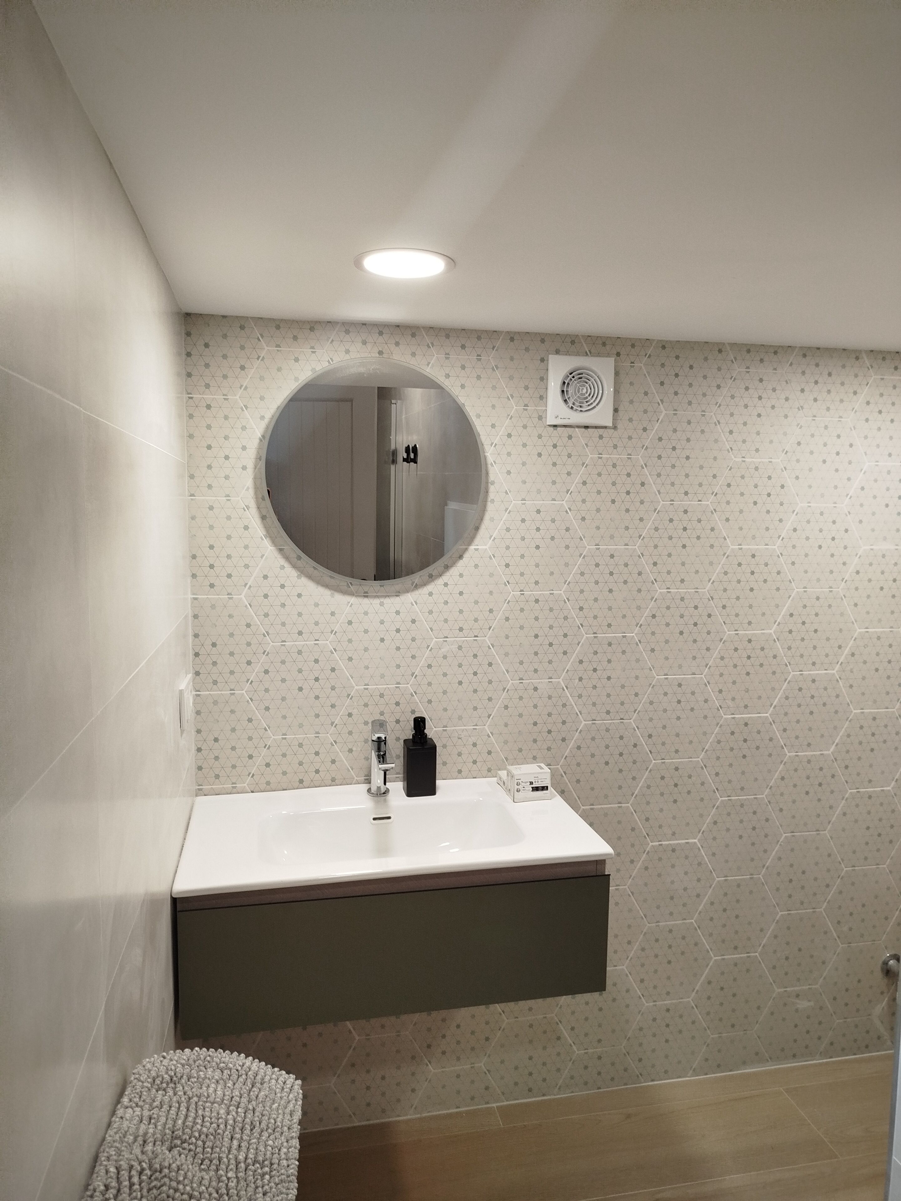 A modern bathroom with a round mirror, a white sink, a black soap dispenser, and a hexagonal patterned wall.