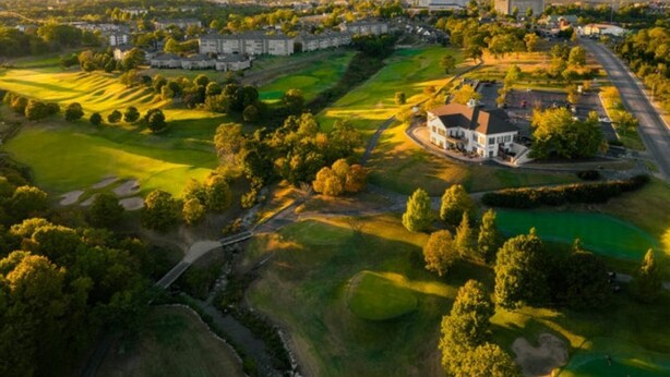 Luftaufnahme eines Golfplatzes mit Klubhaus, umliegenden Gebäuden und einer Straße.