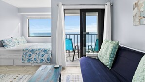 Un dormitorio con cama, sofá, balcón con sillas y vista al mar.