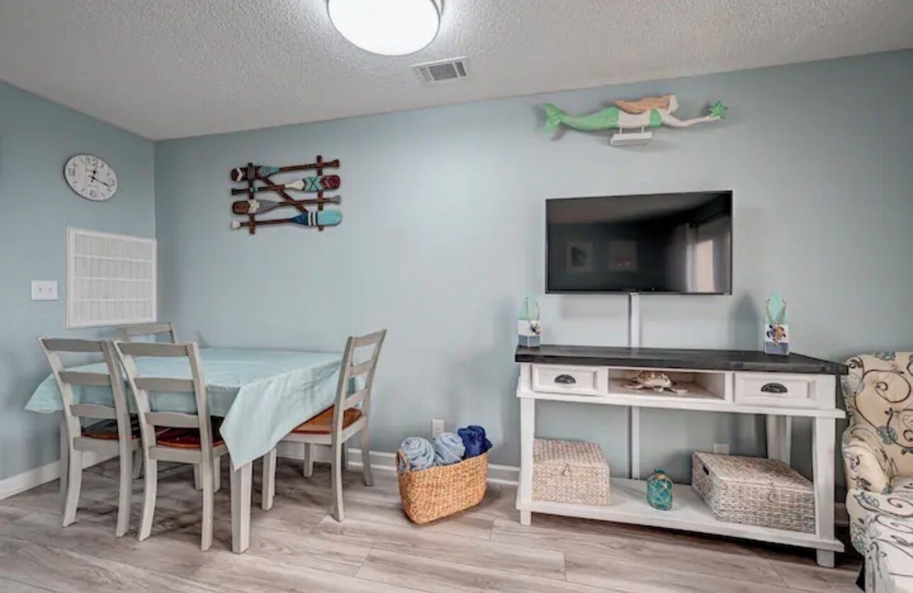 A room with a dining table, chairs, a TV, a desk with drawers, and a wall clock.