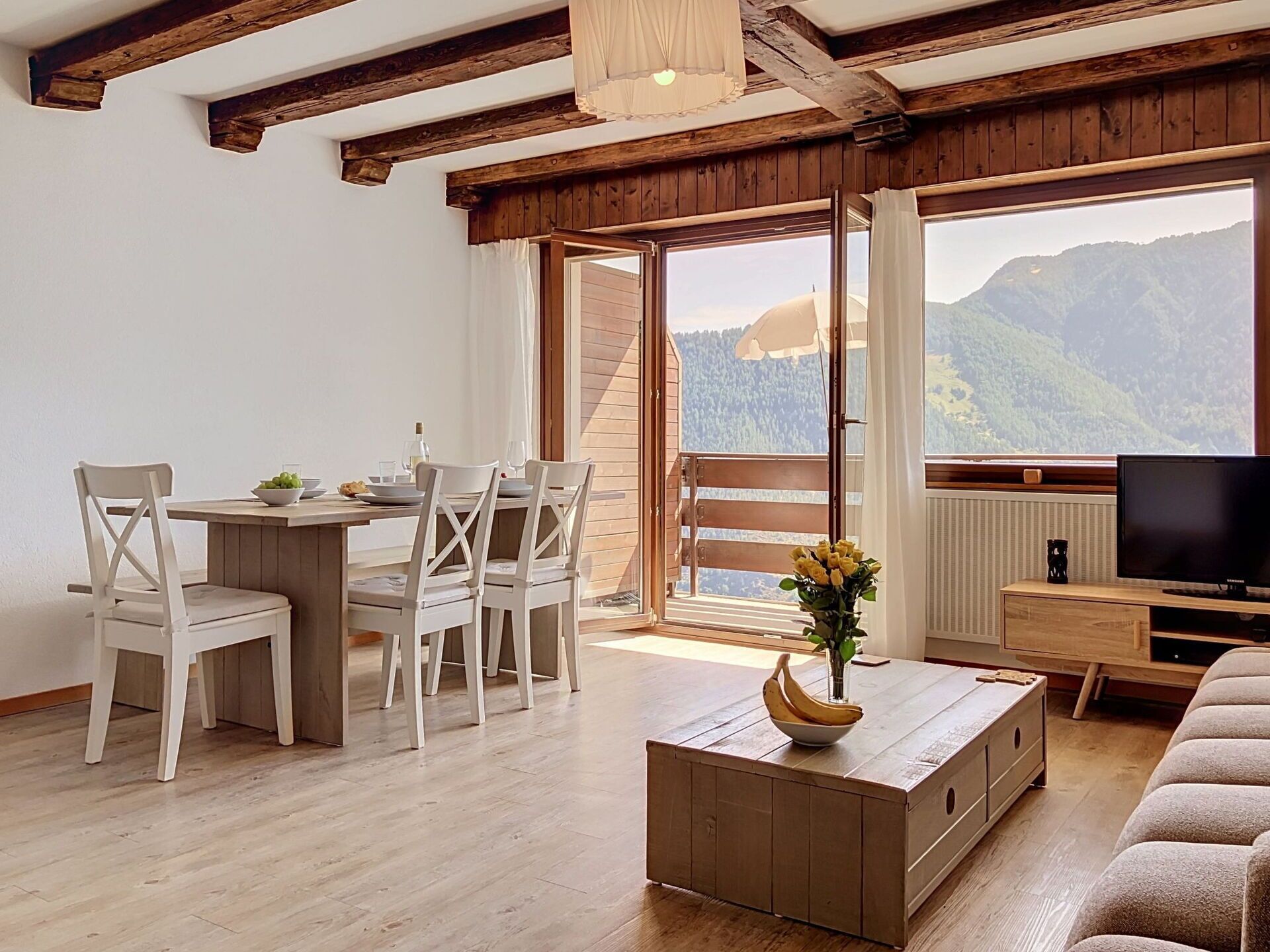 Ein Zimmer mit einem Holztisch, weißen Stühlen, einem Fernseher und Bergblick durch ein großes Fenster.