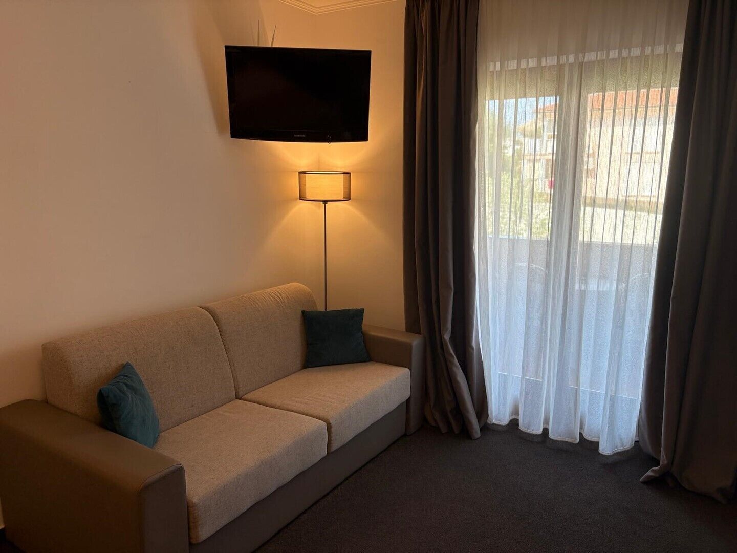 Ein Wohnzimmer mit einer Couch, einem Flachbildfernseher, einer Stehlampe und transparenten Vorhängen.