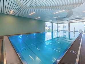 Een groot overdekt zwembad met een modern plafondontwerp, glazen wanden en uitzicht op het buitengebied.