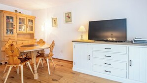 Et spiseområde med et træbord og stole, et skab med glashylder, et fjernsyn på et stativ og en lampe.