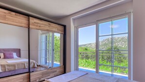 Un dormitorio con una cama grande, cabecera de madera, un espejo y un balcón con vistas a la vegetación y colinas.