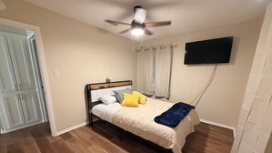 Ein Schlafzimmer mit einem Bett, einem Deckenventilator, einem an der Wand montierten Fernseher und einer Tür.