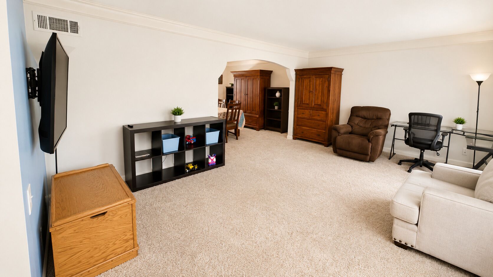 A living room with a TV, a wooden cabinet, a black shelving unit, a brown leather chair, a white sofa, and a desk with a chair.