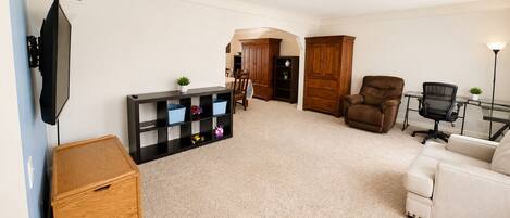A living room with a TV, a wooden cabinet, a black shelving unit, a brown leather chair, a white sofa, and a desk with a chair.