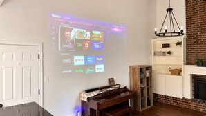 Un soggiorno con un proiettore a muro che mostra l'interfaccia Roku, un pianoforte, un caminetto e un muro in mattoni.