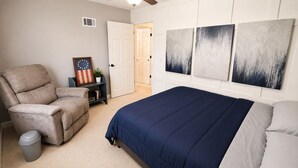 Una camera da letto con un letto, una sedia, un piccolo tavolo con una pianta e opere d'arte alle pareti.