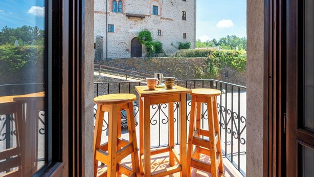 Ein Balkon mit Holzstühlen und einem Tisch, mit Blick auf ein historisches Gebäude mit Rundbogenfenstern und einer Steinmauer.