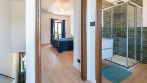 Ein modernes Hotelzimmer mit einem Bett, einem Duschbereich mit Glastüren und einem zur benachbarten Kammer führenden Flur.