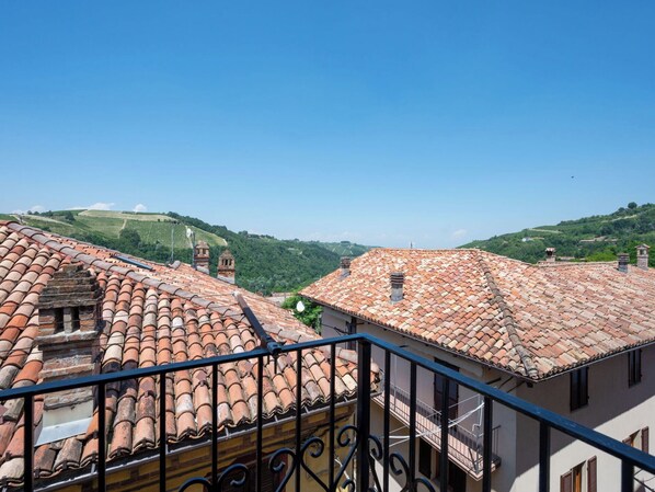 A terrace with red-tiled roofs, a balcony with black railings, and a view of green hills in the background.