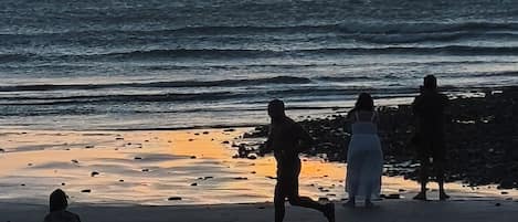 A beach scene with the sun setting over the ocean, people walking along the shore, and a calm sea.