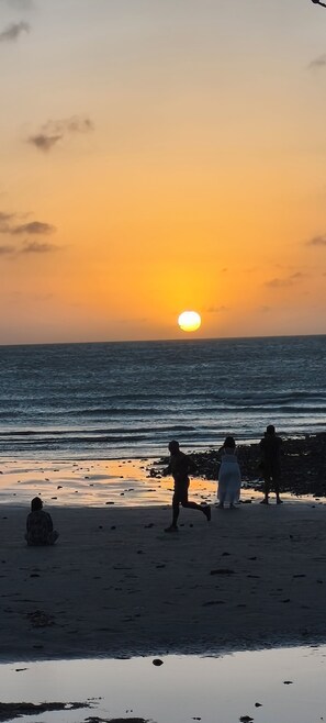 Escena de playa con el sol poniéndose sobre el océano, gente caminando por la orilla y un mar tranquilo.