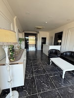 Un pasillo con suelo de mármol, una consola blanca con objetos decorativos, un sofá de cuero negro y un sillón blanco.