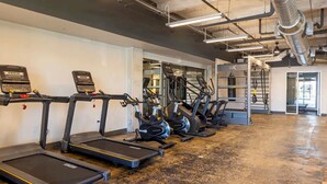 Uma academia com várias esteiras alinhadas em uma parede, equipamentos de exercício e piso de concreto.