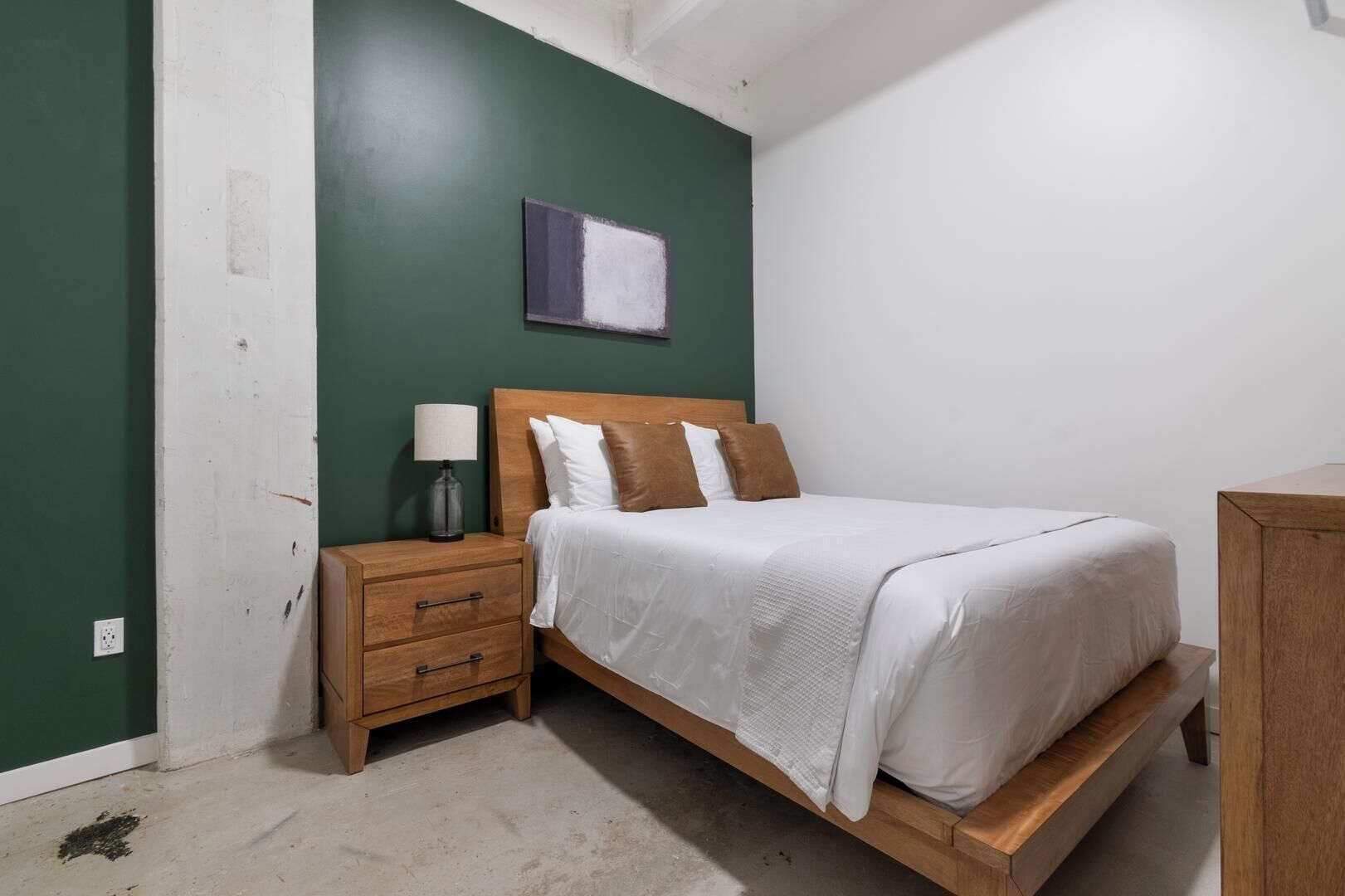 Une chambre à coucher comprenant un lit, une table de chevet en bois, une lampe de chevet et une œuvre d’art accrochée au mur.