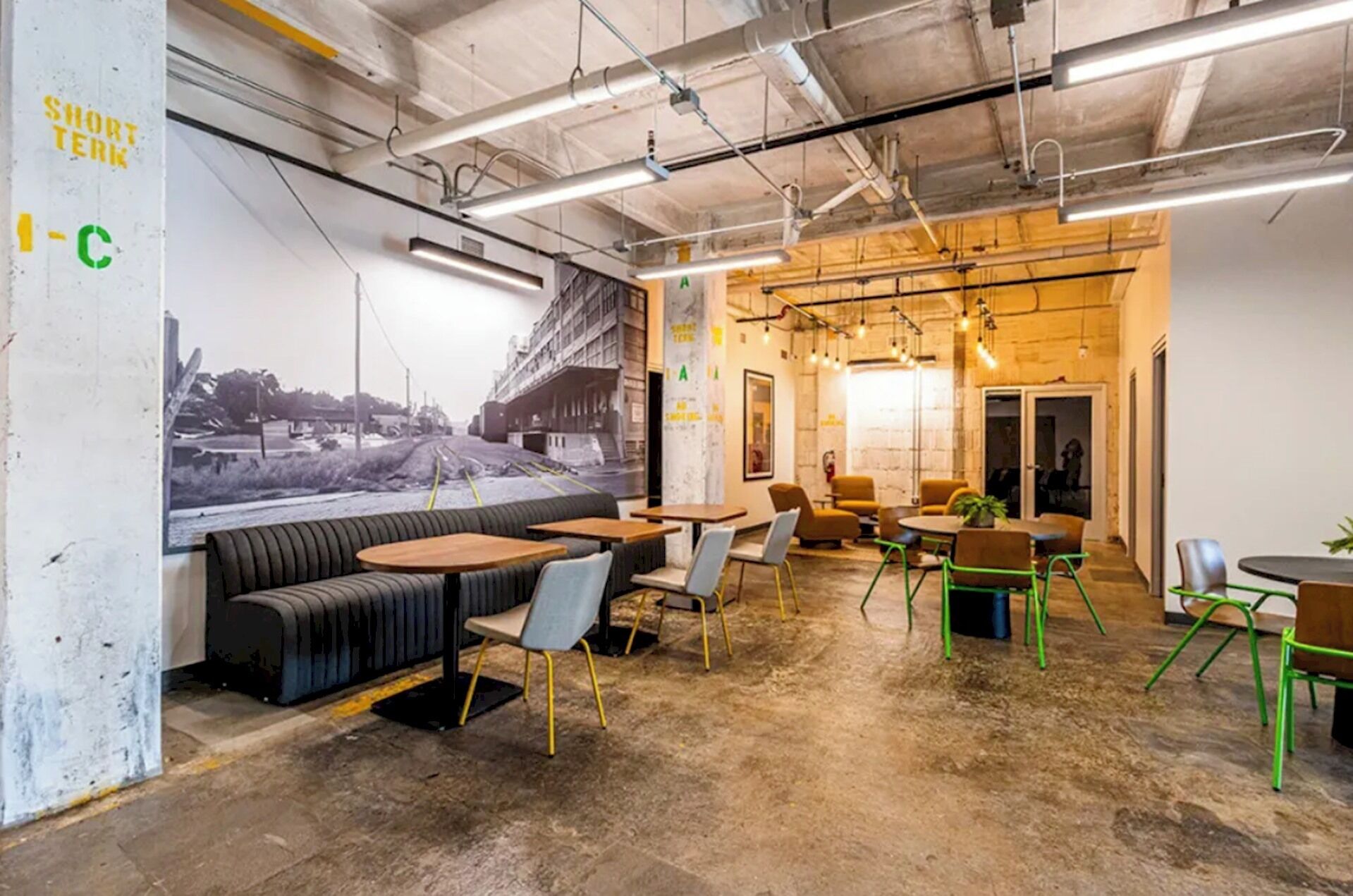 A modern interior with a long bench seating area, tables, and chairs, featuring exposed ceiling pipes and a mural on the wall.