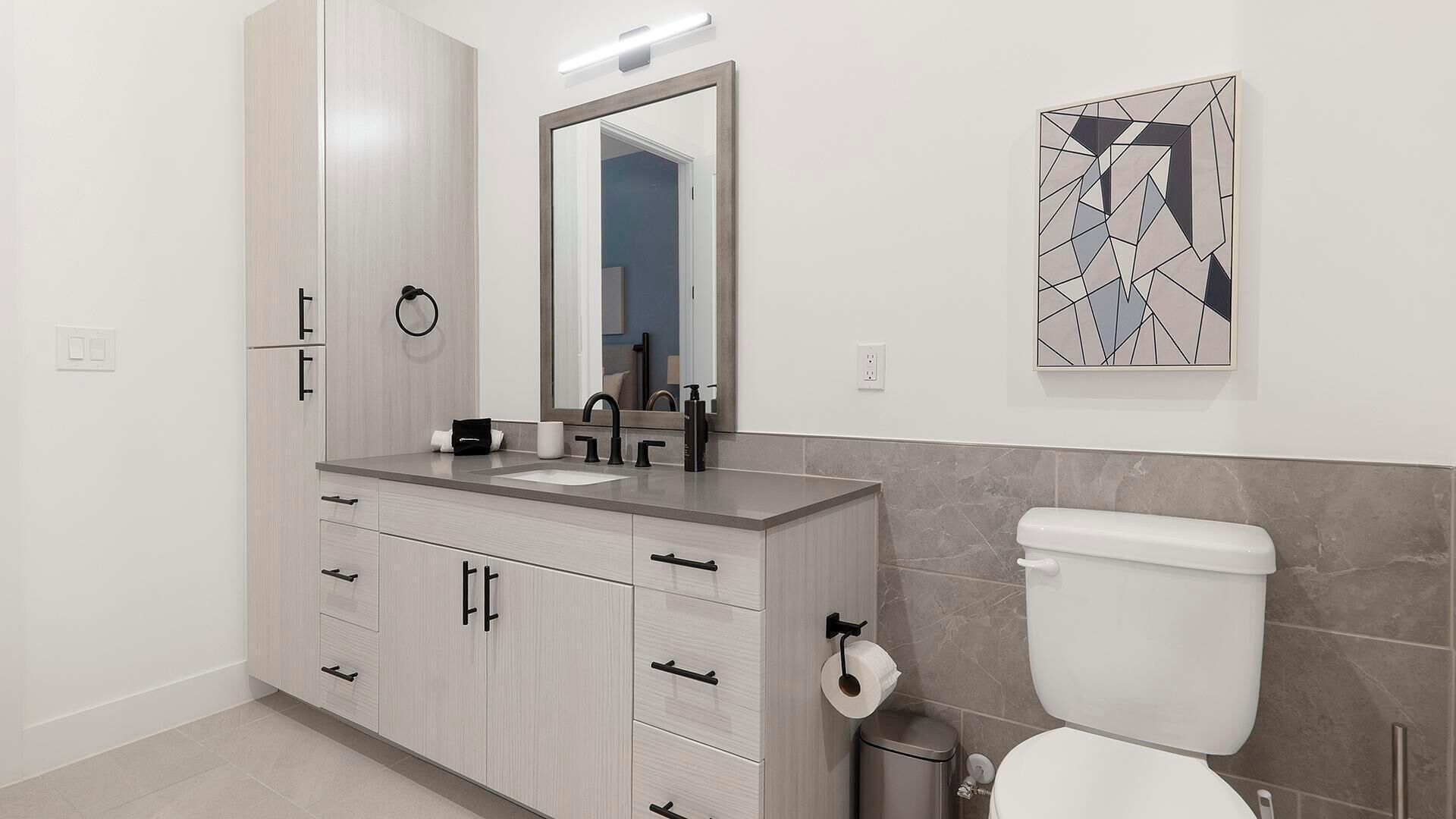Un baño moderno con un mueble de tocador gris, un espejo, un inodoro y un dispensador de papel higiénico en la pared.