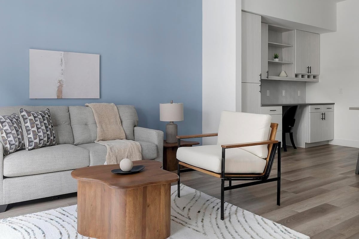 A living room with a grey sofa, patterned pillows, a wooden coffee table, a white armchair, a lamp, and a kitchen area with cabinets.