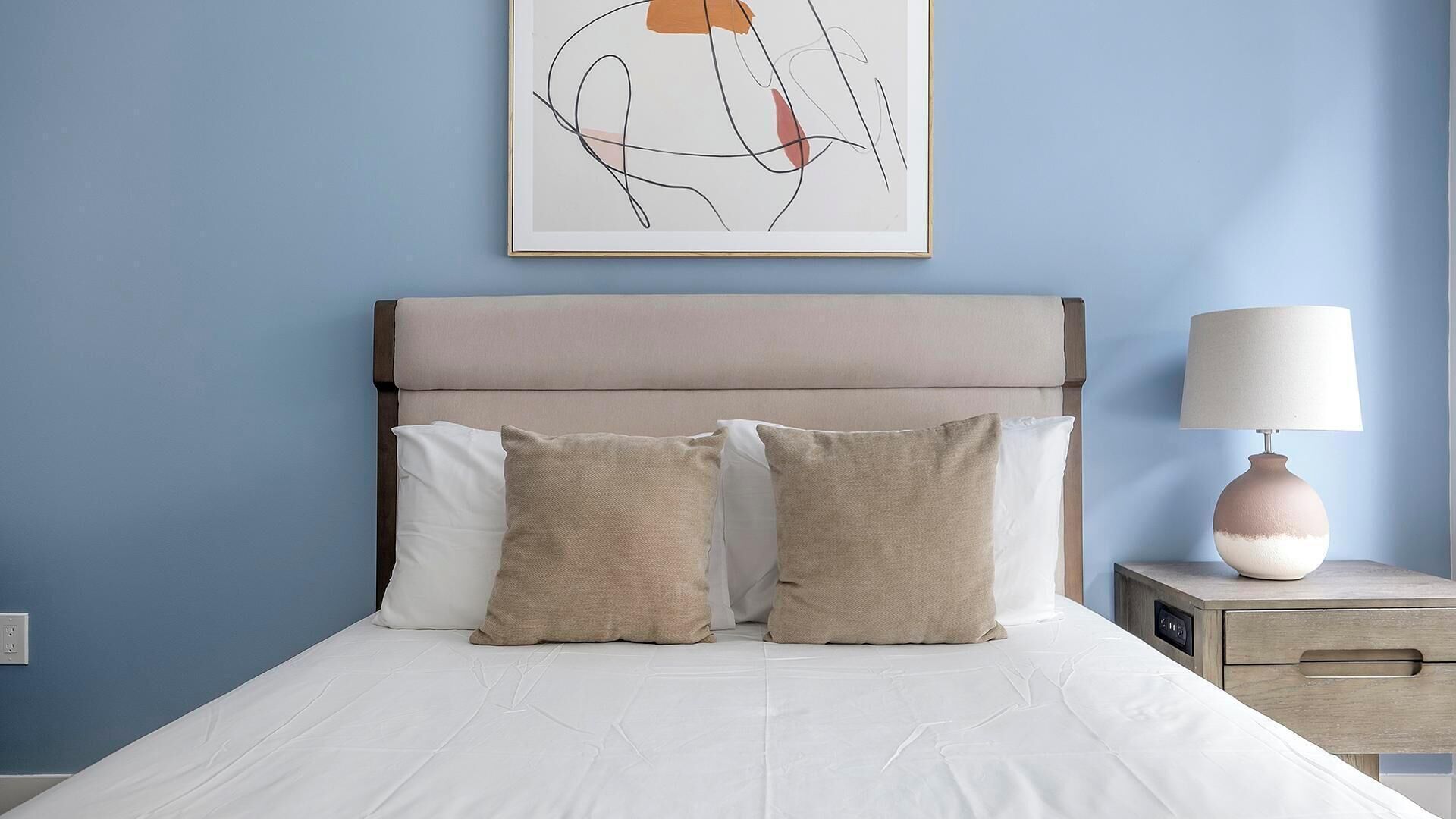 A neatly made bed with white linens and two beige pillows, a bedside table with a lamp, and a wall-mounted abstract art piece.