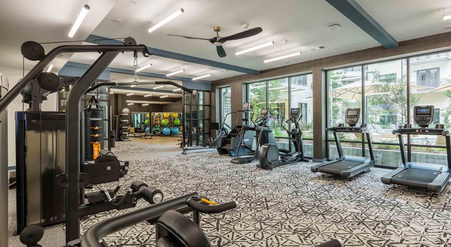 A gym with cardio machines, weight equipment, and large windows offering views of the outdoors.
