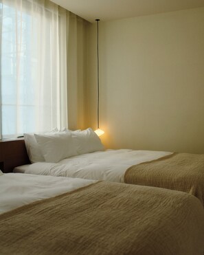 A hotel room with two beds, a nightstand, a lamp, and a window with sheer curtains.