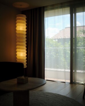 A room with a round table, a lamp, a view through a window with curtains, and a balcony.