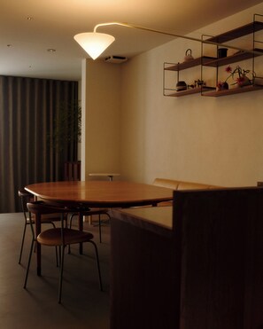 A dining area with a wooden table and chairs, a hanging light fixture, and a shelf with decorative items.