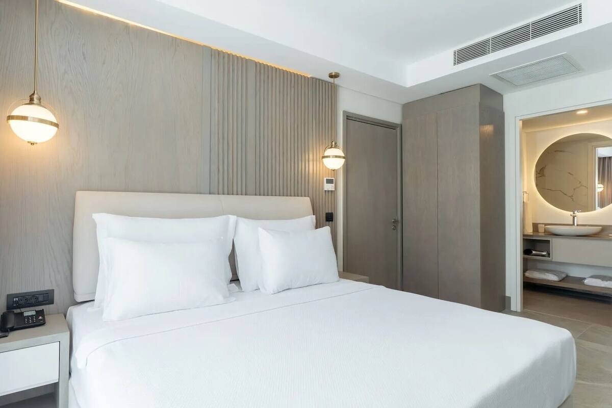 Hotellihuone, jossa on sänky, yöpöytä, lamppu sekä kylpyhuone, jossa on pesuallas ja peili.