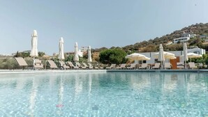 A pool with clear water, surrounded by sun loungers and umbrellas, with buildings and hills in the background.