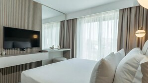 A hotel room with a large bed, a flat-screen TV mounted on a wall, a small desk with a chair, and a large window with curtains.