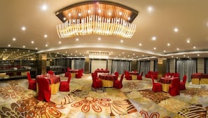 A large banquet hall with round tables and red chairs, a central chandelier, and a patterned carpet. - The India Benares (Varanasi)