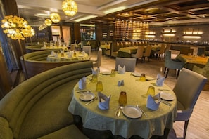 A restaurant with round tables, green upholstered chairs, and a bar area. - The India Benares (Varanasi)
