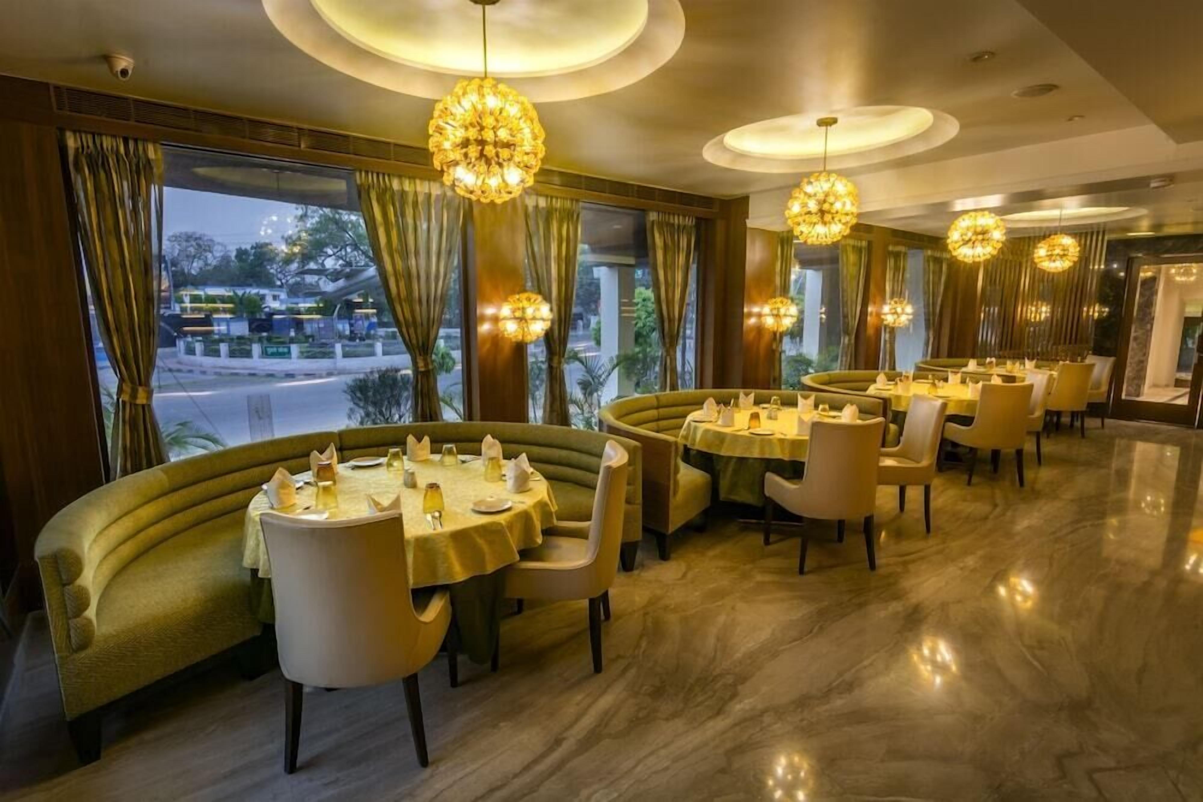 A restaurant with round tables, green cushioned seating, chandeliers, and large windows with curtains.