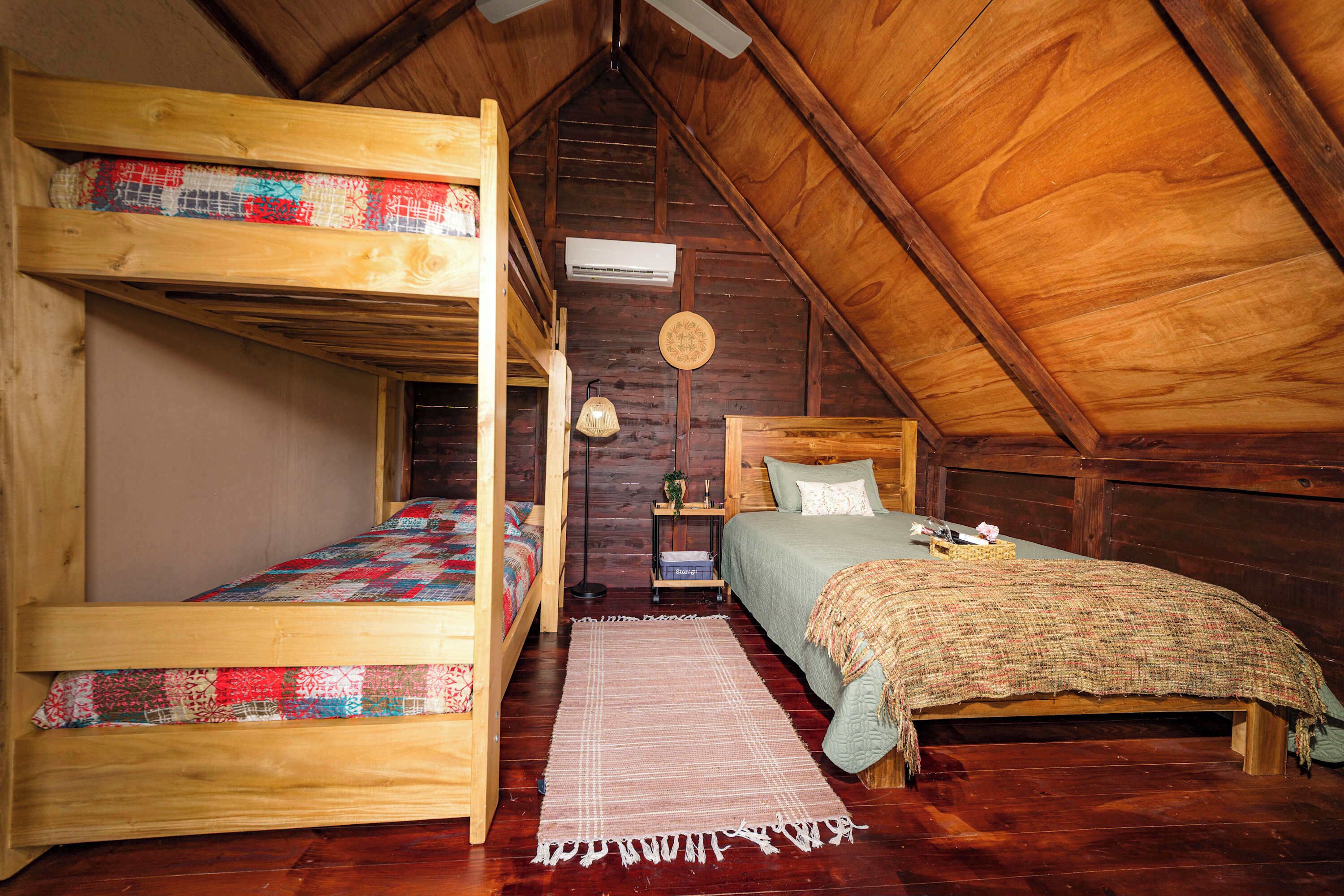 Une pièce avec deux lits superposés, un lit simple, un plafond en bois, une petite table avec une lampe et un tapis sur le sol.