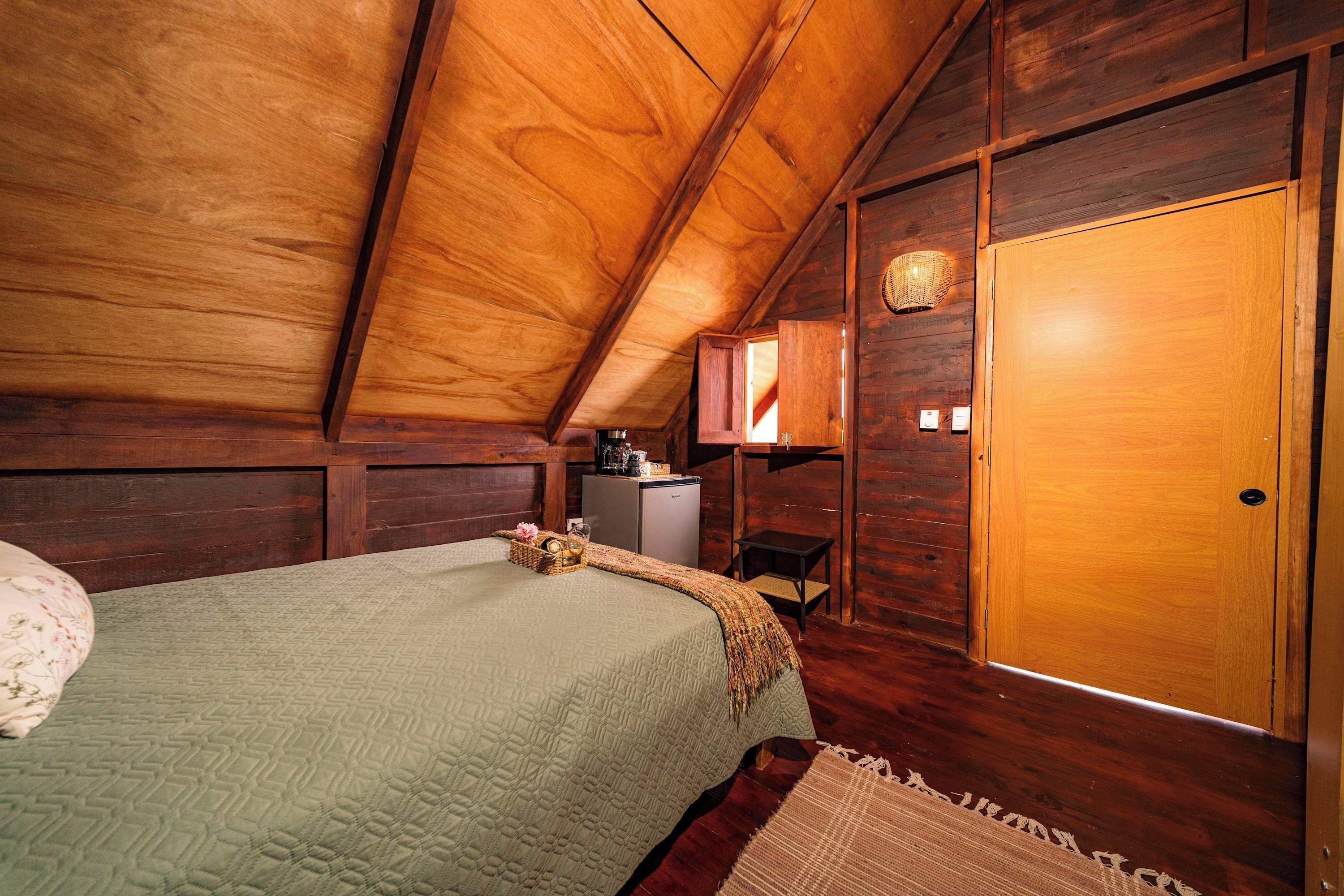 Techo de madera, una cama con colcha verde, un escritorio pequeño con lámpara y una puerta de madera.
