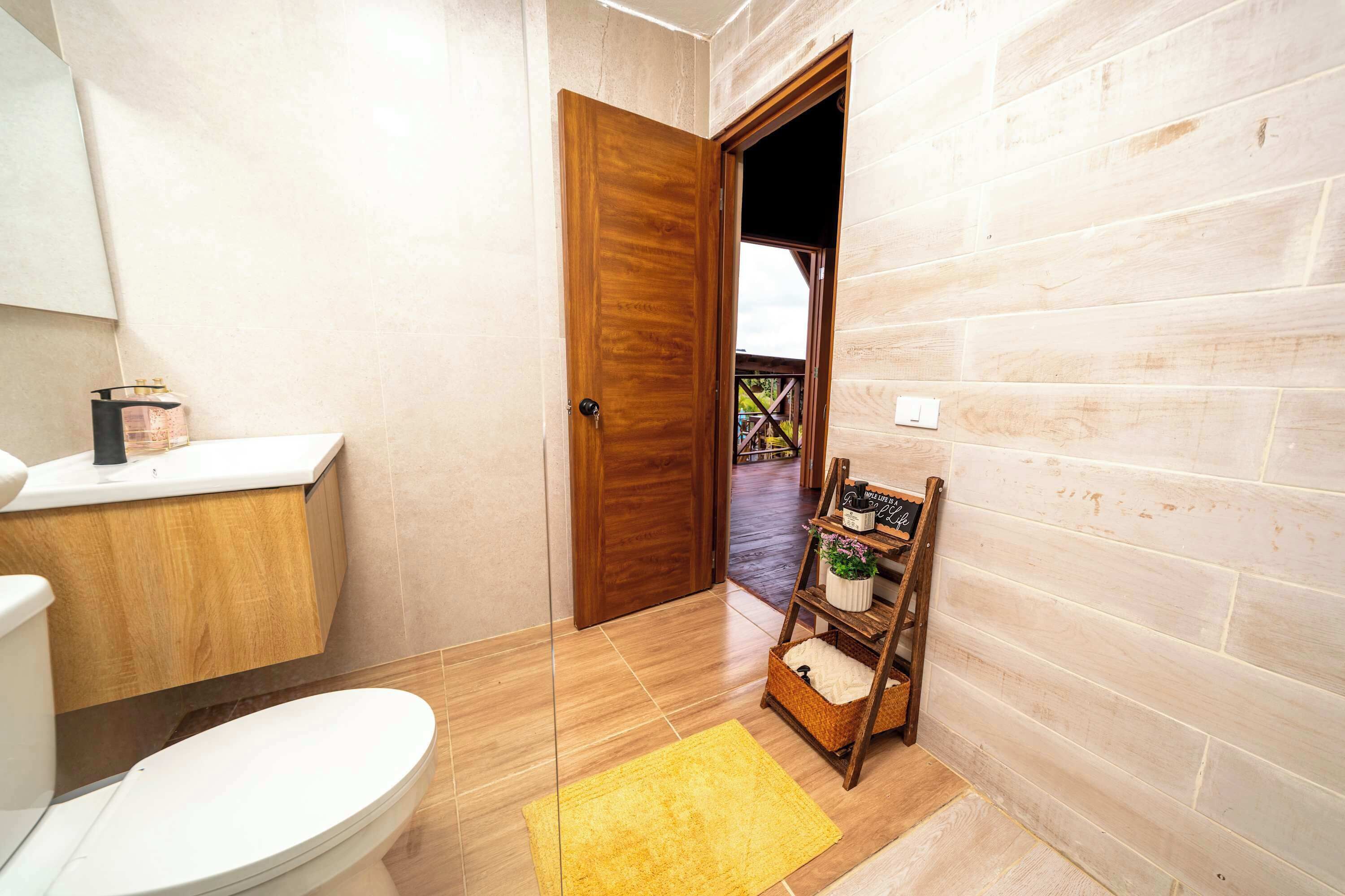 Un baño con puerta de madera, inodoro blanco, alfombra amarilla y estante con una planta.
