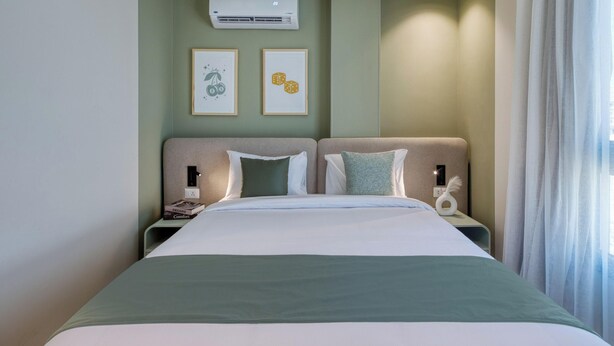 A hotel room with a bed, two bedside tables, a nightstand, a wall-mounted air conditioner, and two framed pictures.
